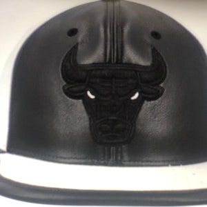 Puede incluir: Una gorra de béisbol de cuero negro con visera blanca y un logotipo de cabeza de toro bordado en negro.