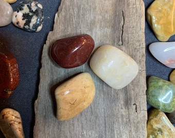 May include: Four smooth, polished stones in various colors, including red, white, and yellow, arranged on a piece of weathered wood.