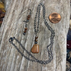 May include: A jewelry set featuring a necklace and earrings. The necklace has a silver chain with a pendant of a light brown stone. The earrings have silver hooks with brown and silver beads. The set is displayed on a wooden surface.