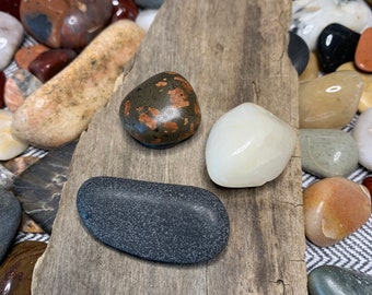 May include: Three smooth, polished stones on a piece of weathered driftwood. The stones are a dark gray, white, and brown with reddish-brown flecks.