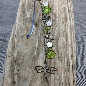 May include: A silver metal bookmark with a dragonfly charm, white flower beads, and green leaf beads. The bookmark is on a wooden surface.