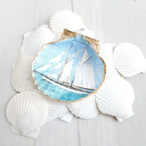 May include: A seashell dish with a painted image of a sailboat on a blue ocean. The seashell is surrounded by other white seashells on a white wooden surface.