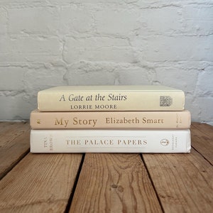 May include: A stack of three books with white covers and gold lettering. The top book is titled "A Gate at the Stairs" by Lorrie Moore. The middle book is titled "My Story" by Elizabeth Smart. The bottom book is titled "The Palace Papers" by Tina Brown.