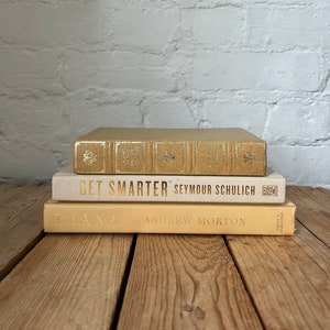 May include: Three hardback books stacked on a wooden surface. The top book is gold with embossed details. The middle book reads "GET SMARTER" in gold lettering. The bottom book is titled "DIANA" in gold text.