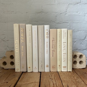May include: A collection of books with white covers and gold lettering. The books are arranged in a row on a wooden surface. The titles of the books include "Jewell", "In the Name", "Something to Tell You", "Reinventing Your Life", "Orr", "Christian Poetry", "Love, Ellen", "A Guide to Being Born", "Light of the World", "Zex in God", and "God"