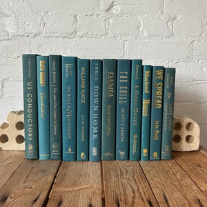 May include: A collection of teal blue hardback books with gold lettering on the spines. The books are arranged vertically on a wooden surface. The titles of the books include "The River Midnight", "Killing Kate", "Down Home", "Damia", "The Chill", "A Good House", "We Spread", and "Lord John".