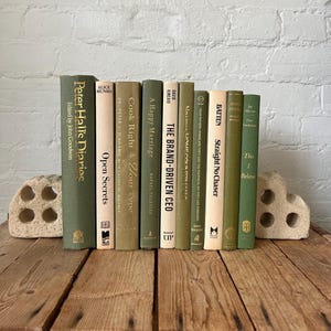 May include: A collection of ten hardback books with spines in shades of olive, sage, and cream, arranged vertically between two textured, light-coloured stone bookends. The titles include Peter Hall's Diaries, Open Secrets, Cook Right 4 Your Type, A Happy Marriage, The Brand-Driven CEO, and Straight No Chaser.