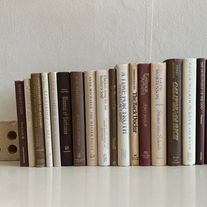 May include: A bookshelf filled with a variety of books, all with brown, cream, and white spines. The books are arranged in a row, with the titles facing outward. The books are all different sizes and shapes, and some have gold lettering on the spines.