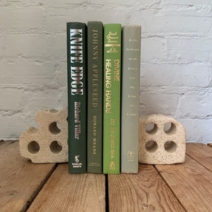 May include: Four green hardback books with gold lettering on the spines are displayed on a wooden surface. The books are held up by a white ceramic bookend. The book titles are "Knife Edge", "Johnny Appleseed", "Divine Healing Hands", and "Don't Let My Baby Do Rodeo".