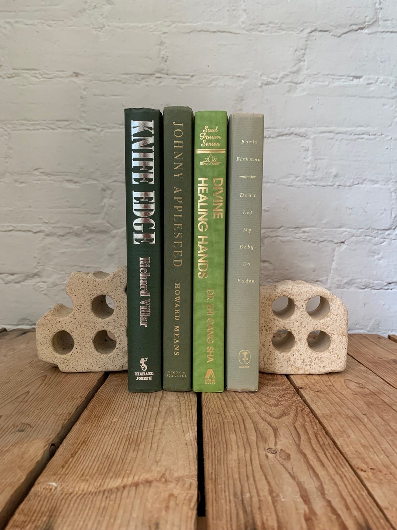 May include: Four green hardback books with gold lettering on the spines are displayed on a wooden surface. The books are held up by a white ceramic bookend. The book titles are "Knife Edge", "Johnny Appleseed", "Divine Healing Hands", and "Don't Let My Baby Do Rodeo".