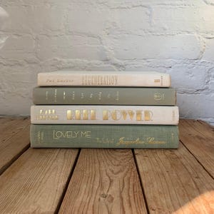 May include: A stack of four hardback books with cloth covers in alternating shades of cream and sage green. The spines feature gold foil lettering including the titles Regeneration, Don't Let My Baby Do Rodeo, Reel Power, and Lovely Me: The Life of Jacqueline Susann.