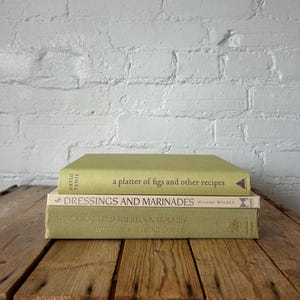 Puede incluir: Tres libros de tapa dura apilados sobre una superficie de madera. El libro superior es de color verde claro con el título "a platter of figs and other recipes". El libro del medio es de color blanquecino con el título "Dressings and Marinades". El libro inferior es de color verde claro con el título "The Annotated Sherlock Holmes."