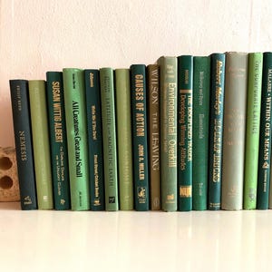 May include: A collection of hardcover books in various shades of green, arranged on a white surface. The spines display titles and authors, creating a visually appealing display. A decorative stone object is on the left.