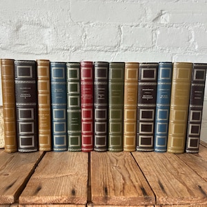May include: A collection of antique books with leather-bound spines in various colors, including tan, blue, green, and red. The books are arranged on a wooden surface, showcasing their decorative details and aged appearance. The spines feature embossed text.