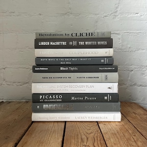 May include: A stack of ten books with grey and white covers. The titles of the books are visible, including "Revolution by Cliché", "The Winter Wives", "Couplehood", "Black Tights", "None to Accompany Me", "The Immune System Recovery Plan", "Picasso: My Grandfather", "Undeniable", and "Chasing Harry Winston".