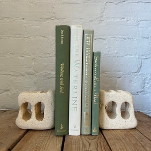 May include: Five books with green covers and gold lettering are displayed on a pair of white stone bookends. The bookends are on a wooden surface. The books are titled "Gardener's Latin", "ETF Investment Strategies", "The Waterline", and "Walking with Jack".