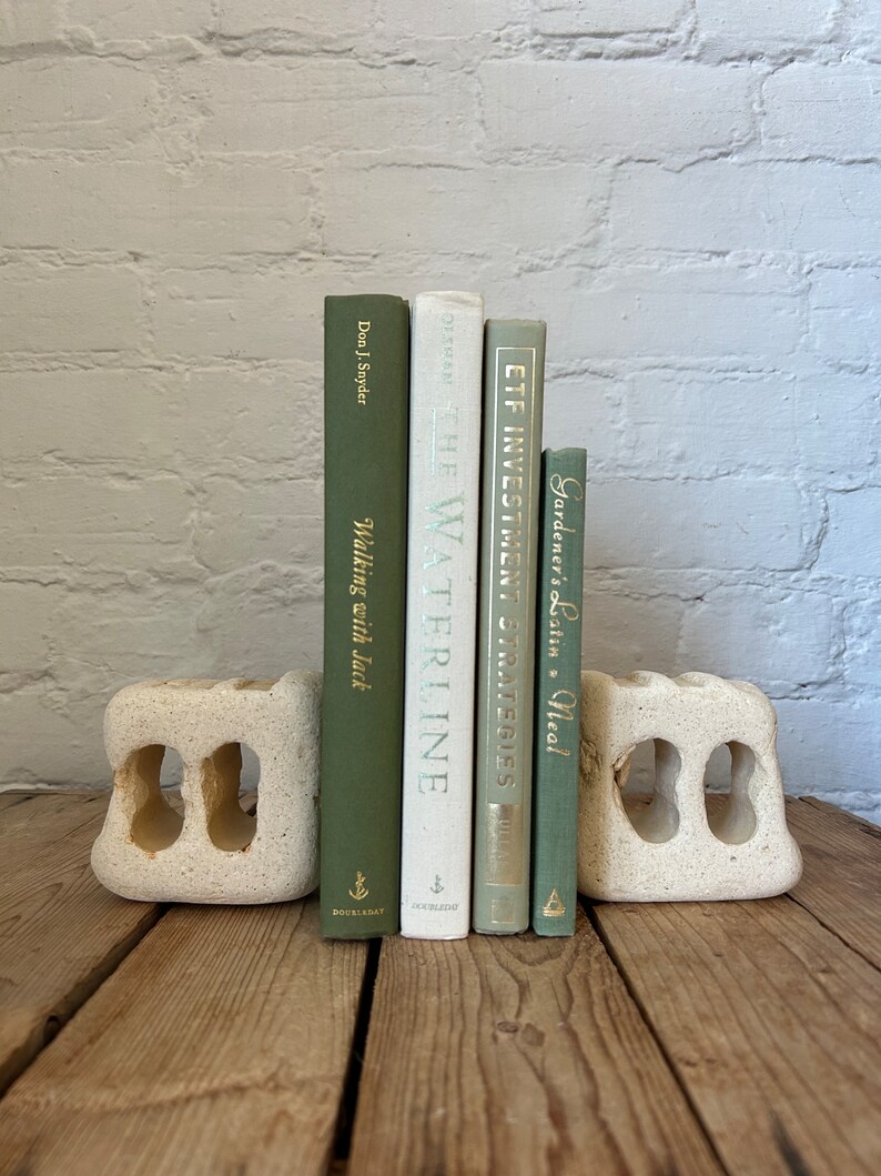 May include: Five books with green covers and gold lettering are displayed on a pair of white stone bookends. The bookends are on a wooden surface. The books are titled "Gardener's Latin", "ETF Investment Strategies", "The Waterline", and "Walking with Jack".
