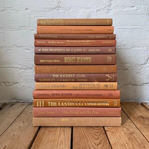May include: A vertical stack of thirteen hardcover books in various shades of brown, tan, and terracotta. The spines feature gold and dark brown lettering displaying titles such as The Hothouse by the East River, Rome Hanks, and The Canadiana Cookbook.