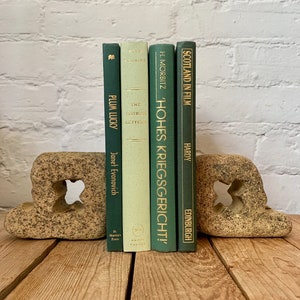 May include: Five green hardback books with gold lettering on the spines are displayed on a pair of stone bookends. The book titles are "Plum Lucky", "The Hatbox Letters", "Hohes Kriegsgericht!", and "Scotland in Film".