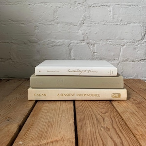 Peut inclure: Une pile de trois livres à couverture rigide aux tons neutres. Le livre du haut est blanc avec le texte "Snowboarding to Nirvana" de Frederick Lenz. Le livre du milieu est gris clair et le livre du bas est crème avec "Gagan: A Sensitive Independence" imprimé en lettres dorées sur le dos.