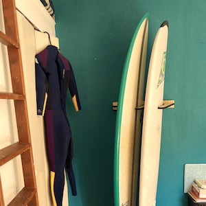 May include: Two surfboards, one white and one green, lean against a teal wall. A wetsuit hangs on a hook to the left. A wooden ladder and a colorful rug are also visible, suggesting a beach or surf theme.