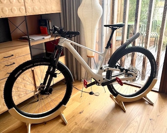 May include: A white mountain bike with black tires and a black seat is displayed indoors on wooden stands. The bike has red pedals and a black front fender. The background includes a wooden cabinet, a paper lantern, and a window.