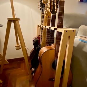 以下が含まれることがあります： 木製のギター用スタンドに、アコースティックギターとエレキギターが複数本立てかけられています。ギターは天然木仕上げで、一列に並んでいます。フロアランプとソファが背景にあります。