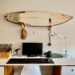 May include: A surfboard mounted on a wall above a desk. The surfboard is white with black and orange accents and some graffiti. A desk with a computer, keyboard, and other accessories is below the surfboard.