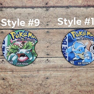 May include: Two round, colorful, printed, cardboard coasters with a Pokemon theme. The left coaster features a green version of the Pokemon character, Venusaur, with the text "Gotta catch 'em all!" and "Green Version". The right coaster features a blue version of the Pokemon character, Blastoise, with the text "Gotta catch 'em all!" and "Blue Version".