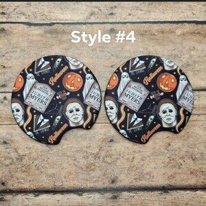 May include: Two black car coasters with a Halloween theme. The coasters feature images of a jack-o'-lantern, a ghost, and the face of Michael Myers. The text "Halloween", "Judith Myers", and "Dr. Loomis" are also visible.