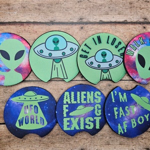 May include: Set of six round green and blue drink coasters with alien and UFO designs. The coasters feature phrases like "Get in Loser, We're Going to the UFO World", "Aliens Exist", and "I'm Fast AF Boy".