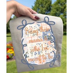 May include: A light gray sweatshirt with the text "Being their mama is who I'm meant to be" in orange script. The text is inside a floral patchwork design with blue, peach, and white flowers.