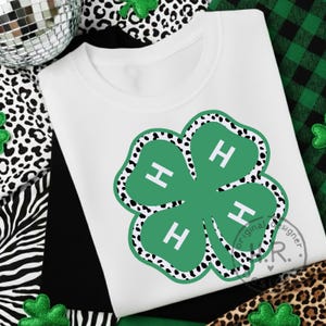 May include: White t-shirt featuring a green four-leaf clover design with the letter "H" in each leaf. The clover has a black and white spotted border. The shirt is surrounded by decorative elements including a disco ball and shamrock accents.