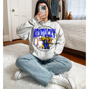 May include: A light gray hoodie with a graphic design featuring the text "Kentucky Wildcats" in blue, a baseball, and a yellow and blue wildcat logo. The hoodie has a drawstring hood and a kangaroo pocket.