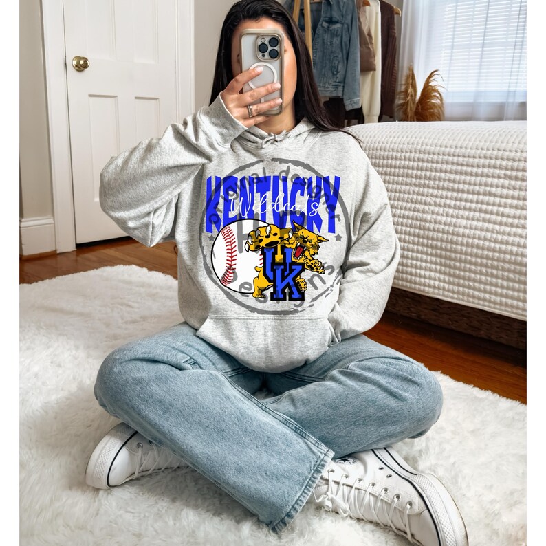 May include: A light gray hoodie with a graphic design featuring the text "Kentucky Wildcats" in blue, a baseball, and a yellow and blue wildcat logo. The hoodie has a drawstring hood and a kangaroo pocket.