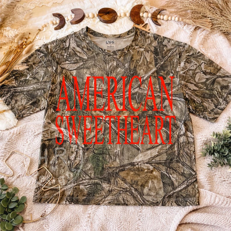 May include: A short-sleeved camouflage t-shirt with the words "AMERICAN SWEETHEART" in large red letters across the front. The shirt has a crew neck and is made of a lightweight material. The background is a soft, neutral color.
