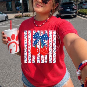Puede incluir: Camiseta roja con un diseño patriótico con rayas rojas y blancas, estrellas azules y un lazo con cerezas. La camiseta tiene cuello redondo y mangas cortas. Una taza blanca con un logotipo rojo se sostiene en la mano izquierda.