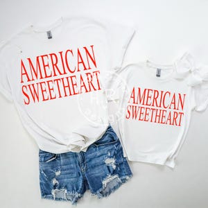 May include: White t-shirts with the text "AMERICAN SWEETHEART" in red. One shirt is paired with denim shorts. A smaller shirt with the same text and a white bow is also visible. The shirts are a casual style.