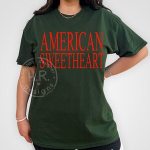 May include: Dark green t-shirt with the words "AMERICAN SWEETHEART" in red block letters. The shirt has short sleeves and a relaxed fit. The design is centered on the front of the shirt.
