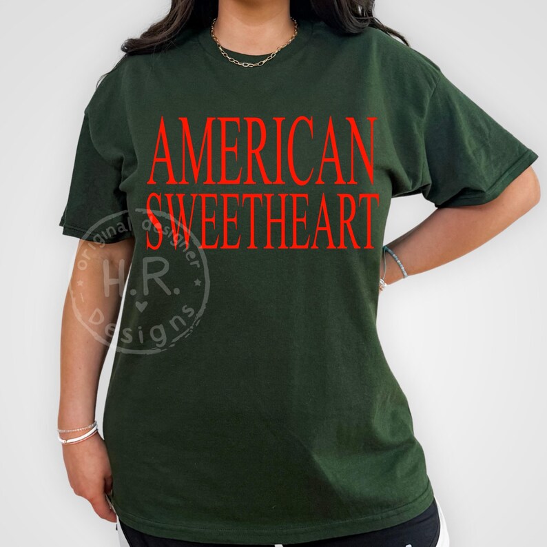 May include: Dark green t-shirt with the words "AMERICAN SWEETHEART" in red block letters. The shirt has short sleeves and a relaxed fit. The design is centered on the front of the shirt.