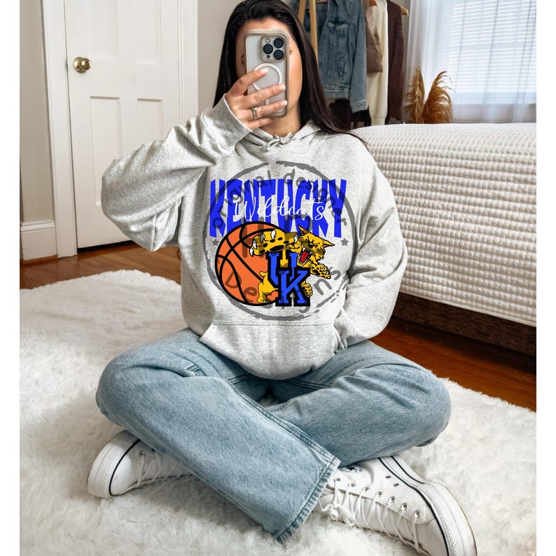 May include: A light gray hoodie with a graphic design featuring the word "Kentucky" in blue, a basketball, and a wildcat. The hoodie has a drawstring hood and a kangaroo pocket. The person is wearing light blue jeans and white sneakers.