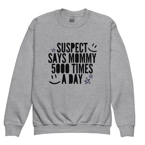Puede incluir: Una sudadera gris con el texto "SUSPECT SAYS MOMMY 5000 TIMES A DAY" impreso en negro. El texto está rodeado de estrellas y una carita sonriente.