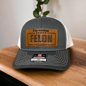 May include: A gray and white trucker hat with a brown leather patch that says "I'm voting FELON - 2.24" with a small American flag design. The hat has a white mesh back and a Richardson 112 adjustable strap.