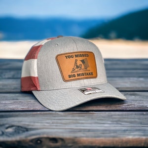 May include: A gray and red trucker hat with a brown leather patch that says "You Missed Big Mistake". The patch features a black and white illustration of a person holding a flag.