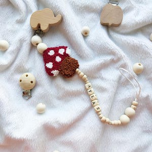 Könnte beinhalten: Ein Baby-Schnullerclip mit einem rot-weißen Pilzdesign, Holzperlen und dem Namen "ATLAS" in Holzbuchstaben. Der Clip enthält hölzerne Elefanten- und Apfelformen.