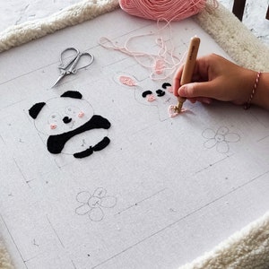 Op de afbeelding: Een close-up van een witte stof met zwarte en roze borduursels van een panda, konijn en bloem. Een hand gebruikt een houten borduurwerktool om het konijn te naaien.