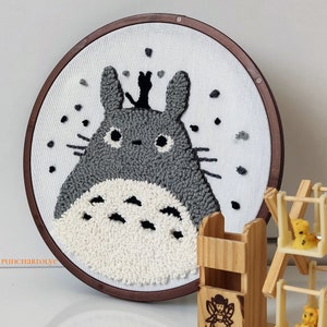 May include: A gray and white embroidered Totoro character on a white canvas in a brown wooden embroidery hoop.