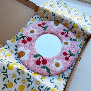 May include: A pink round mirror with a white floral and cherry design. The mirror is surrounded by a white border and is set against a white background with a lemon pattern.