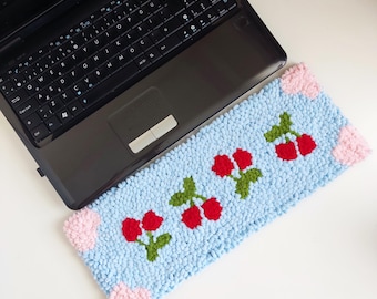 May include: A light blue, rectangular laptop mat with a textured surface. The mat features a repeating pattern of red cherries with green leaves. Pink hearts are in each corner. A black laptop is partially visible.