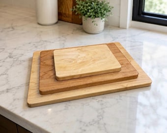 May include: A stack of three rectangular wooden cutting boards of varying sizes and wood tones, ranging from light to medium brown, arranged in a tiered formation on a white marble surface with visible grey veining throughout the stone.